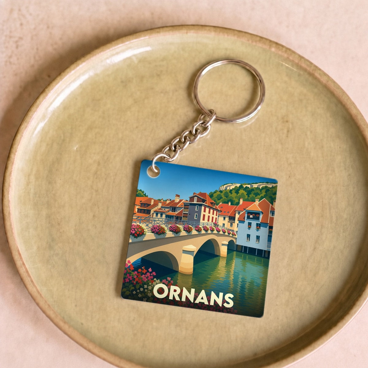 Porte-clés souvenir de la ville d'Ornans avec une illustration stylisée de son pont fleuri et de ses maisons au bord de l'eau.