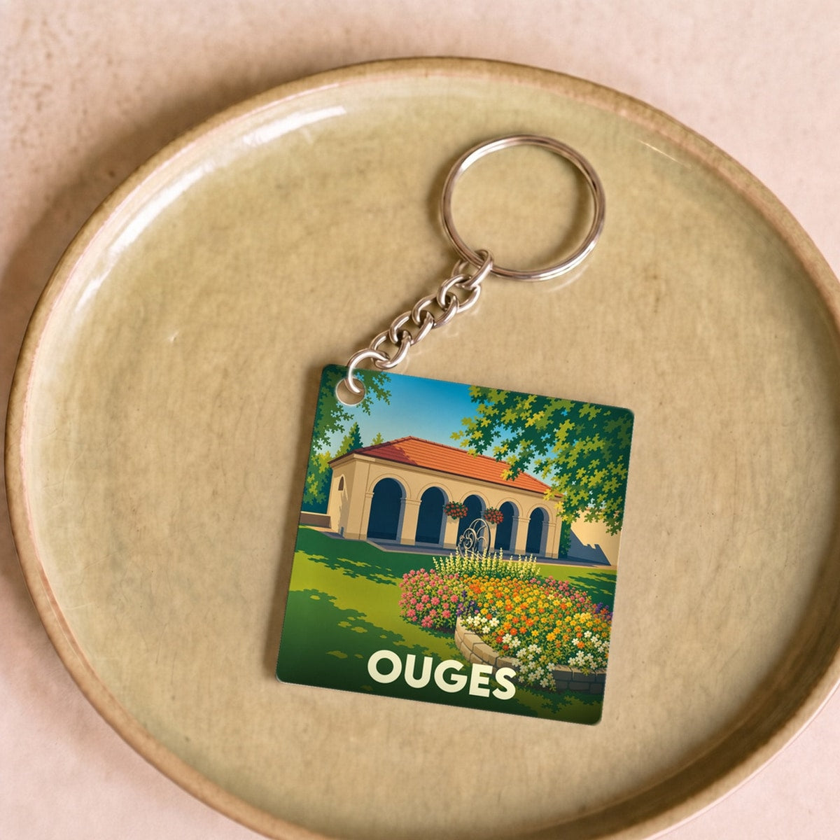 Porte-clés souvenir de la ville de Ouges avec une illustration stylisée d'un bâtiment à arcades et de jardins fleuris.