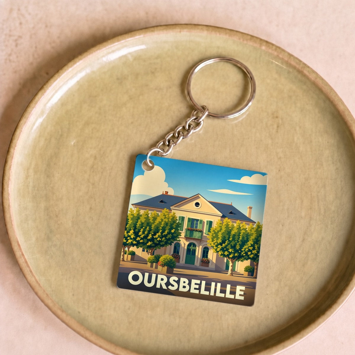 Porte-clés souvenir de la ville d'Oursbelille avec une illustration stylisée d'une maison de maître entourée d'arbres.