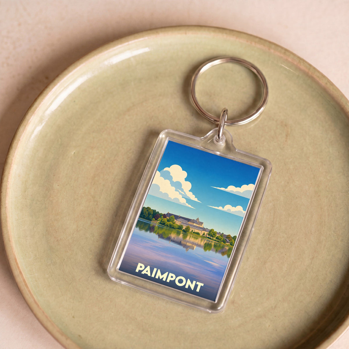 Porte-clés souvenir de la ville de Paimpont avec une illustration stylisée de son abbaye au bord de l'eau.