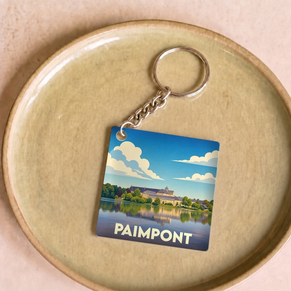 Porte-clés souvenir de la ville de Paimpont avec une illustration stylisée de son abbaye au bord de l'eau.