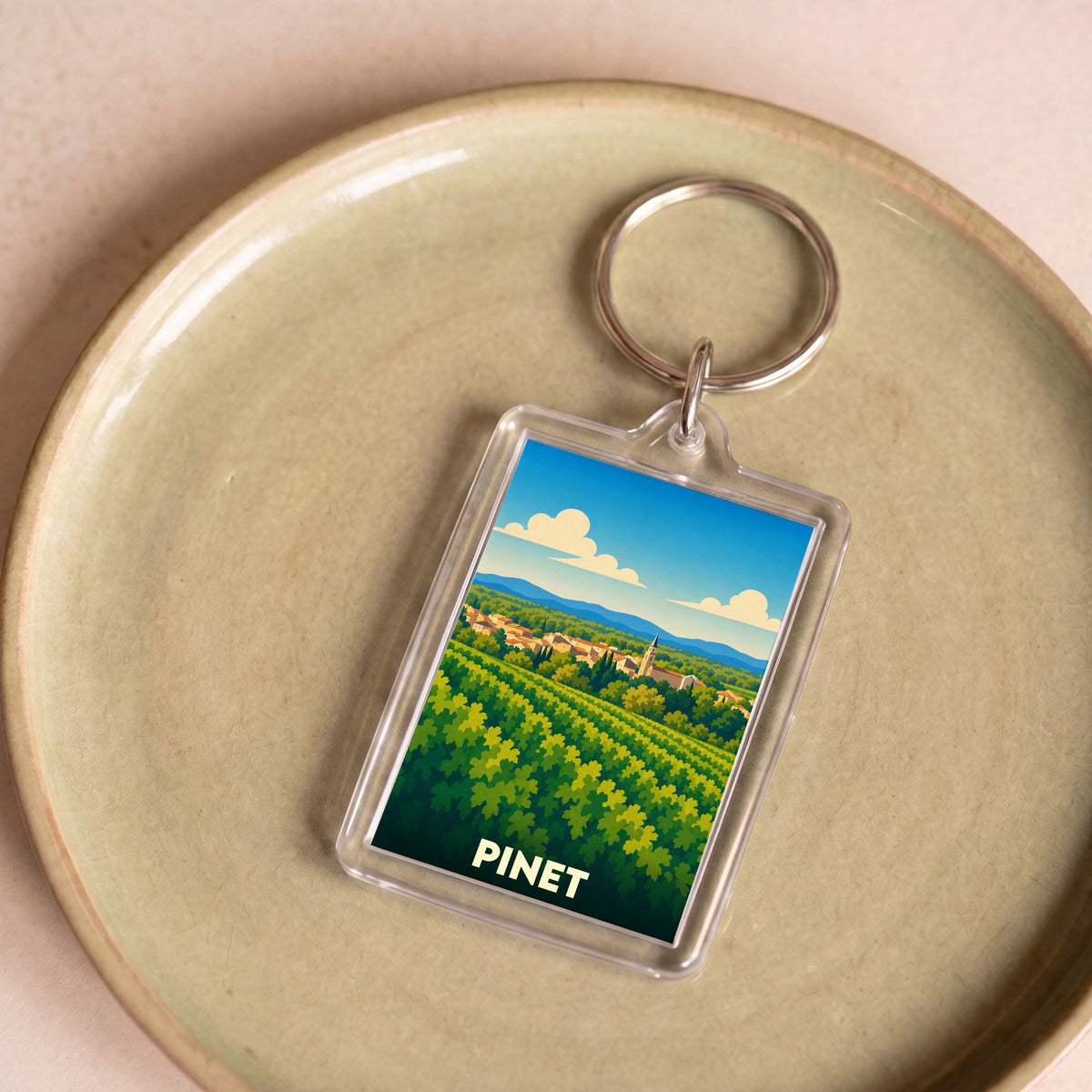 Porte-clés souvenir de la ville de Pinet avec une illustration stylisée de vignobles et du village.