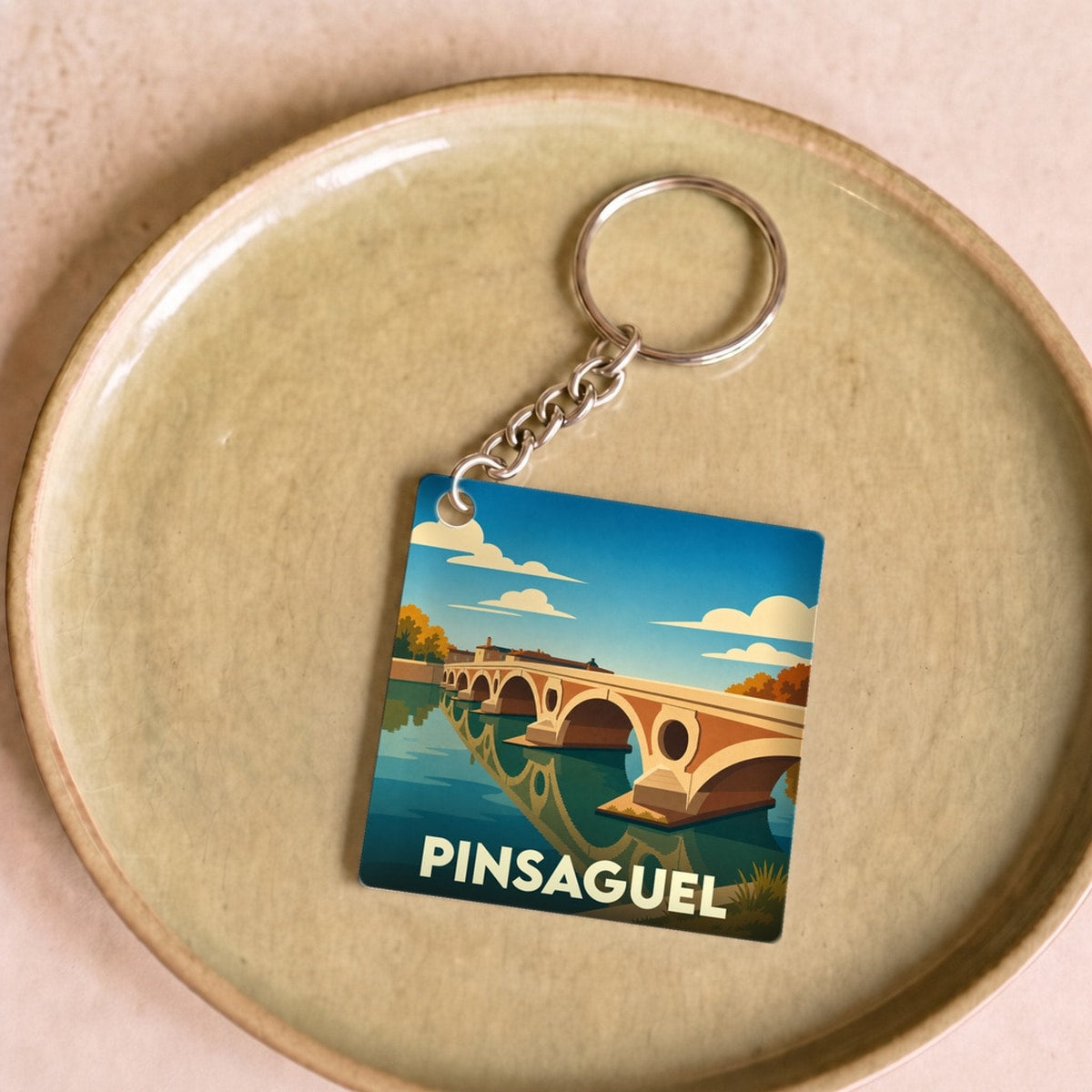 Porte-clés souvenir de la ville de Pinsaguel avec une illustration stylisée d'un pont enjambant une rivière.