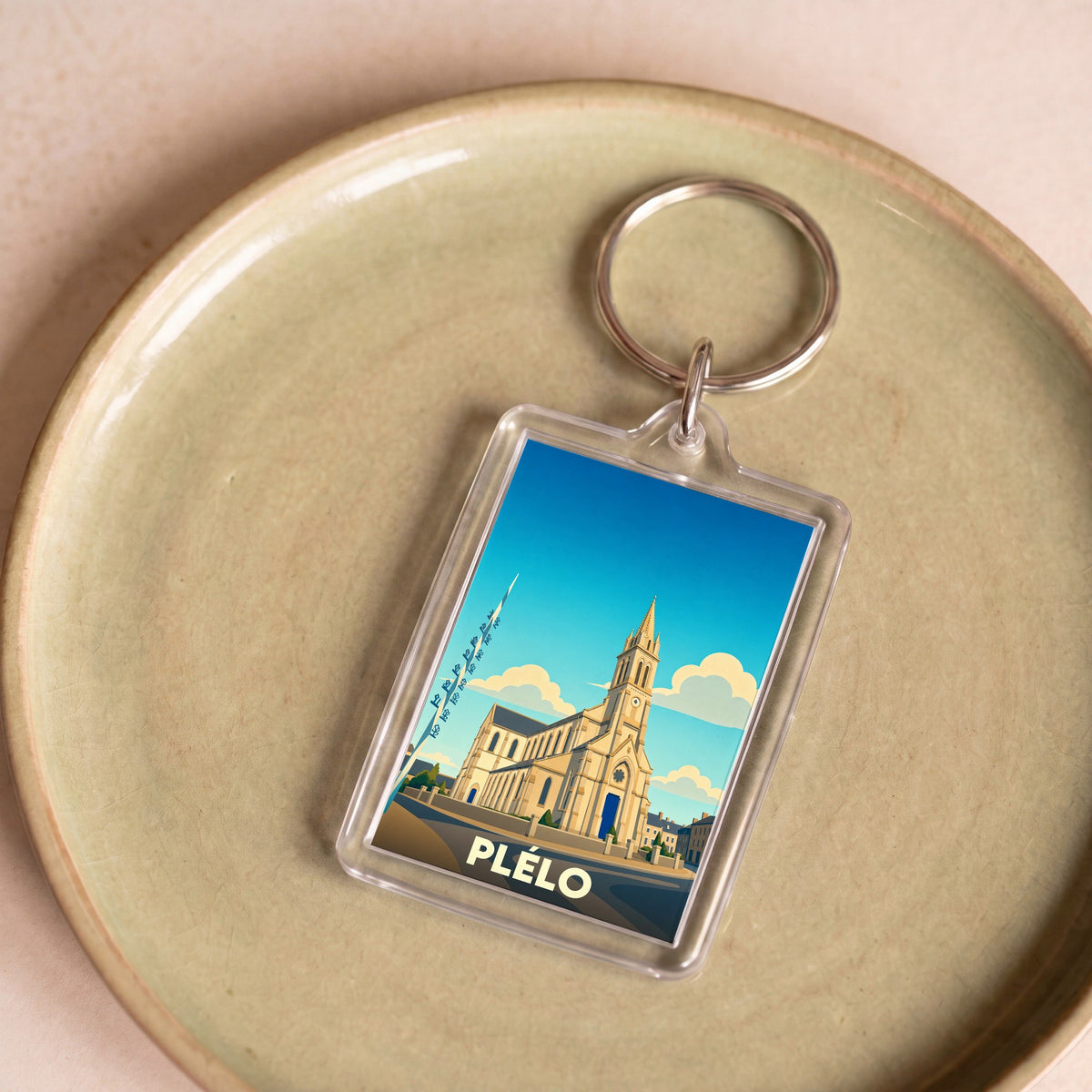 Porte-clés souvenir de la ville de Plélo avec une illustration stylisée de son église sous un ciel bleu.