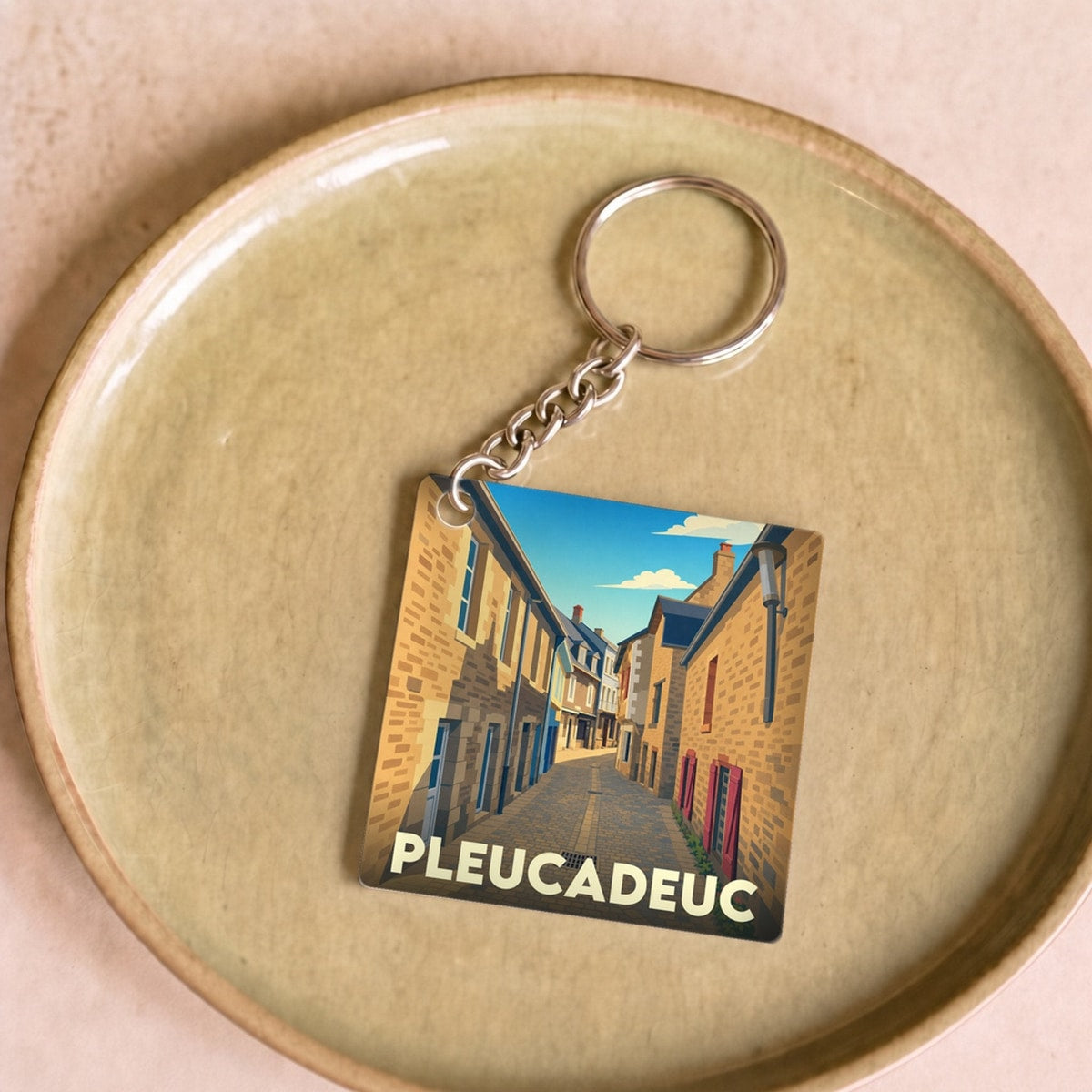 Porte-clés souvenir de la ville de Pleucadeuc avec une illustration stylisée d'une rue pittoresque.
