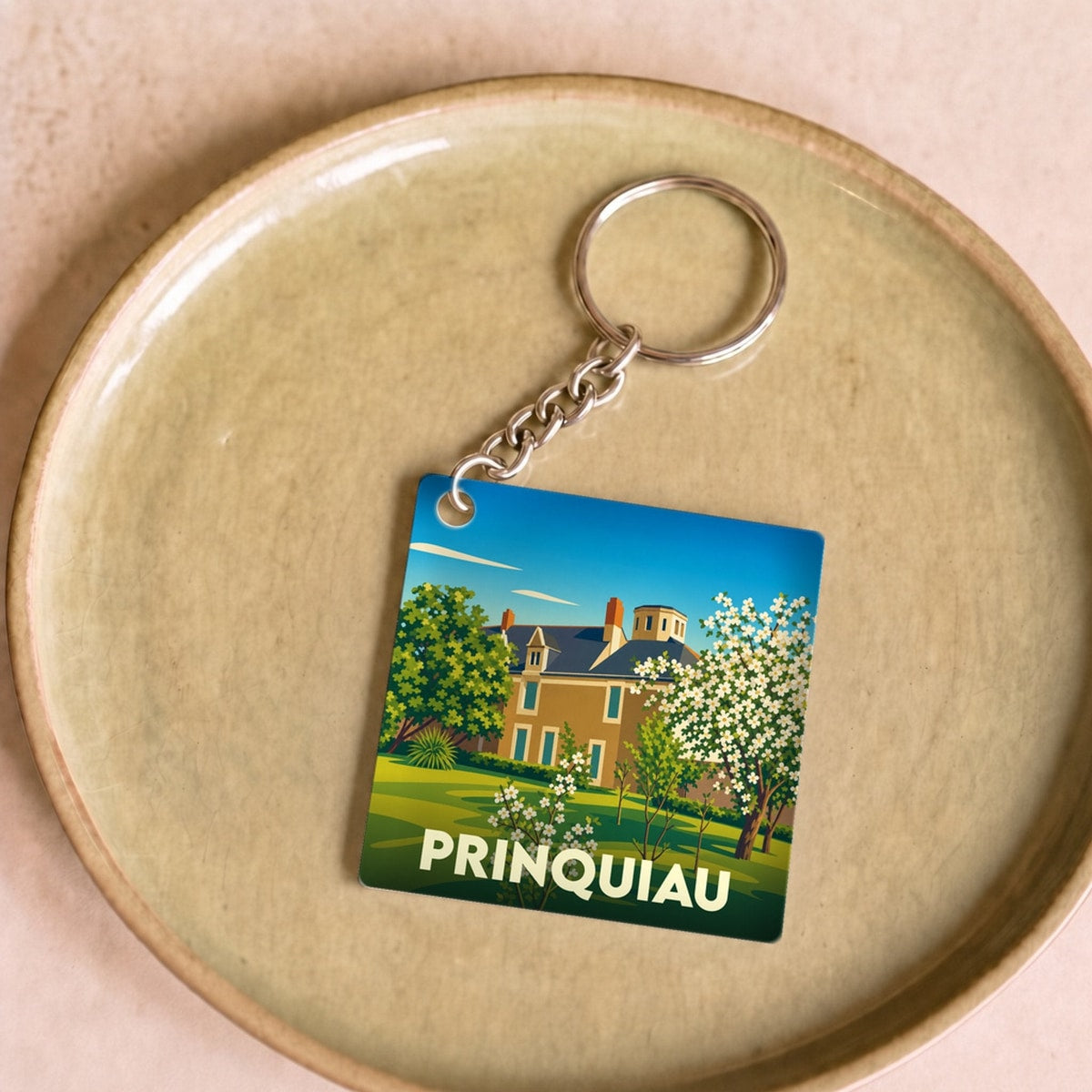 Porte-clés souvenir de la ville de Prinquiau avec une illustration stylisée d'une demeure et de son jardin.