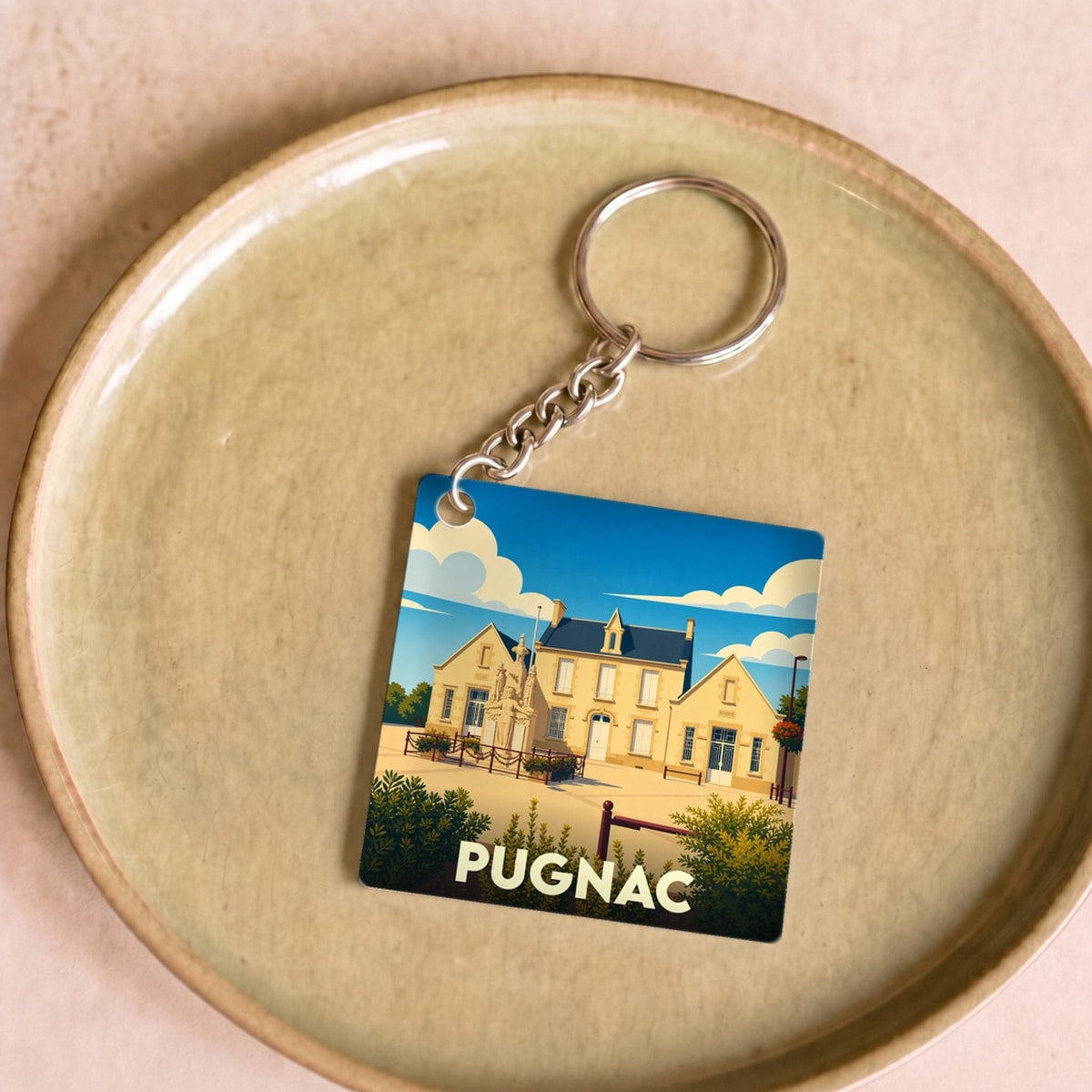 Porte-clés souvenir de la ville de Pugnac avec une illustration stylisée de sa mairie sous un ciel bleu.