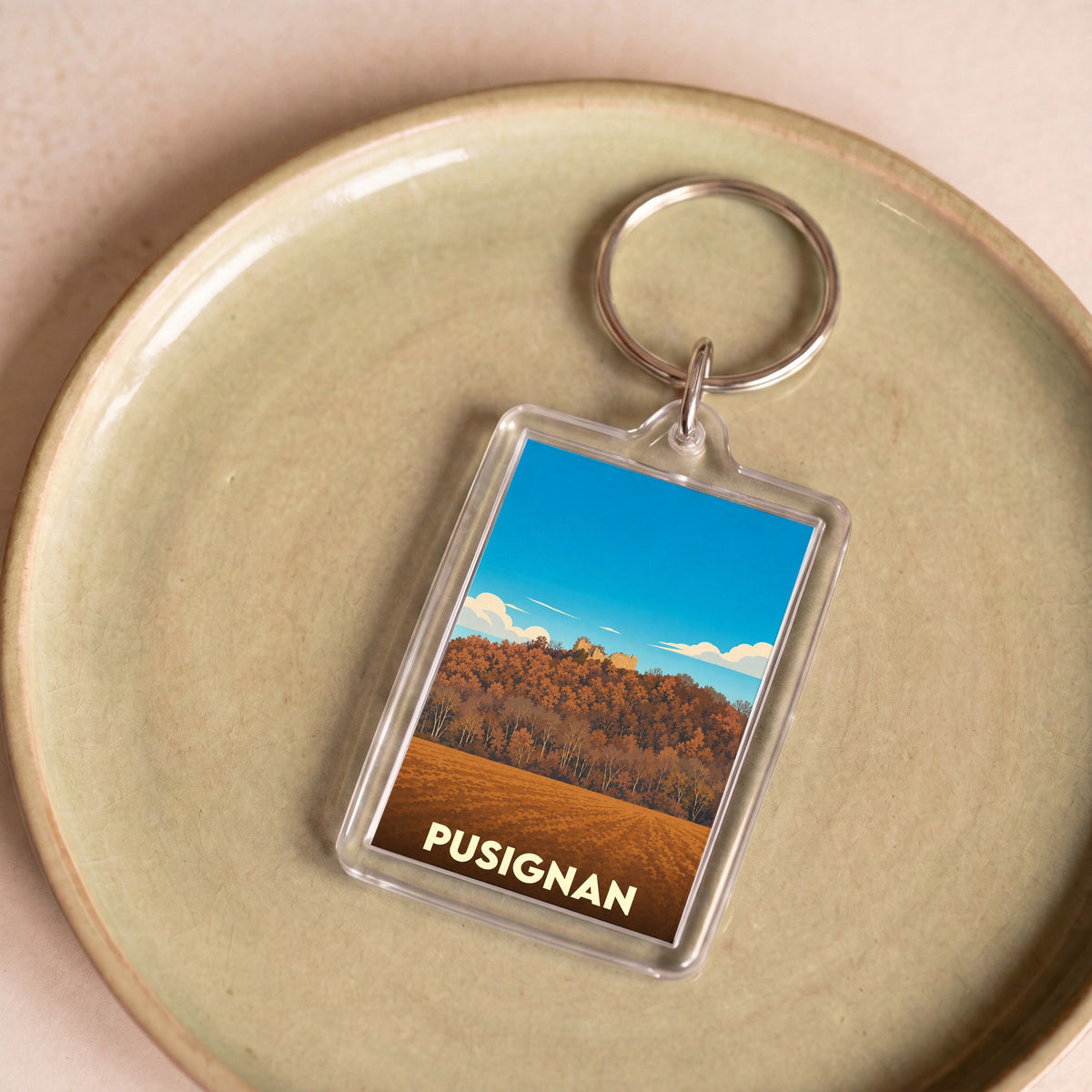 Porte-clés souvenir de la ville de Pusignan avec une illustration stylisée d'un paysage automnal.