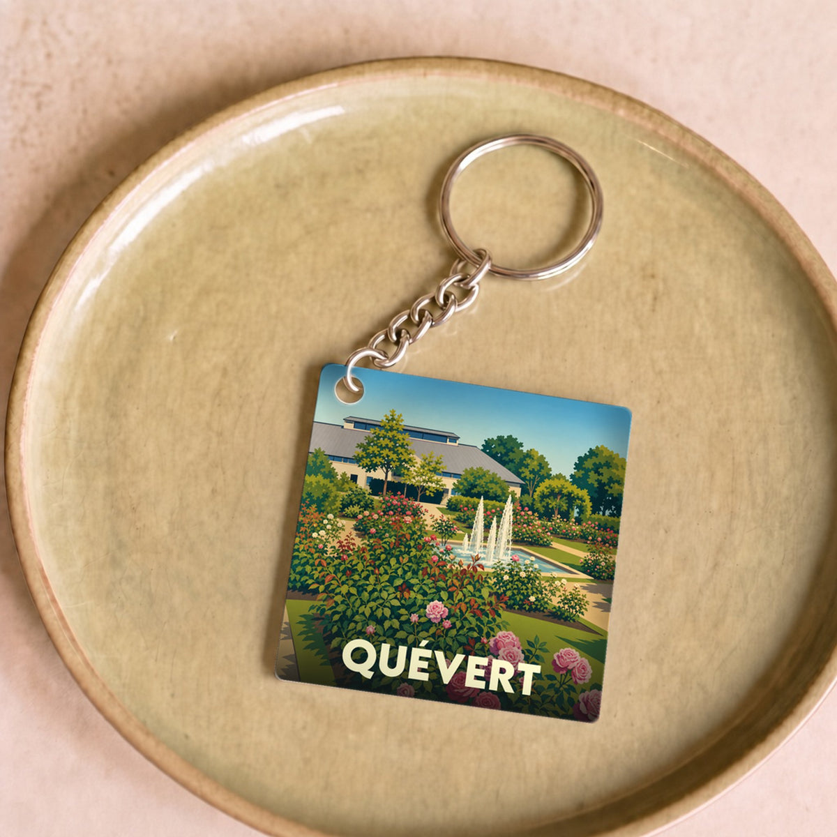 Porte-clés souvenir de la ville de Quévert avec une illustration stylisée d'un jardin fleuri et de ses fontaines.