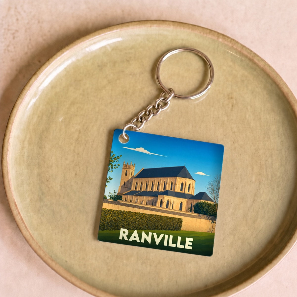 Porte-clés souvenir de la ville de Ranville avec une illustration stylisée de son église sous un ciel bleu.