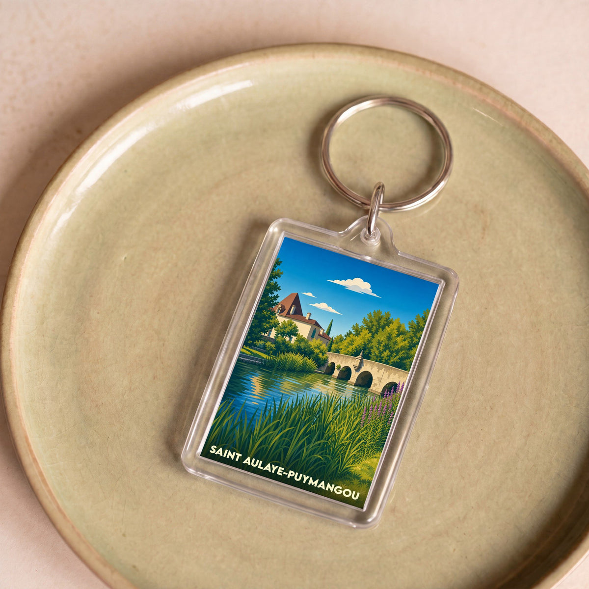 Porte-clés souvenir de la ville de Saint Aulaye-Puymangou avec une illustration stylisée d'un pont et d'un manoir au bord de l'eau.