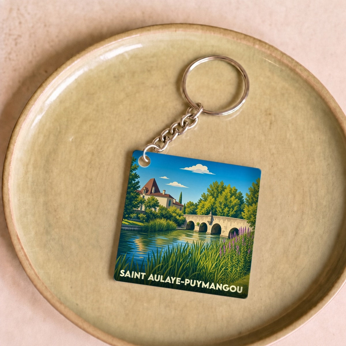 Porte-clés souvenir de la ville de Saint Aulaye-Puymangou avec une illustration stylisée d'un pont et d'un manoir au bord de l'eau.