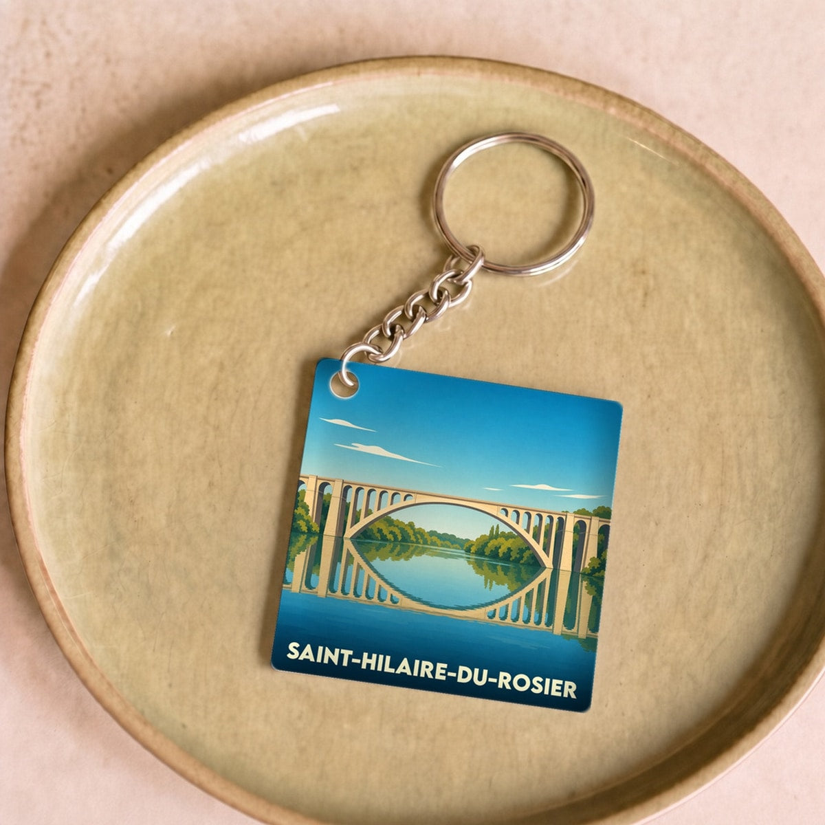 Porte-clés souvenir de la ville de Saint-Hilaire-du-Rosier avec une illustration stylisée de son pont se reflétant dans l'eau.