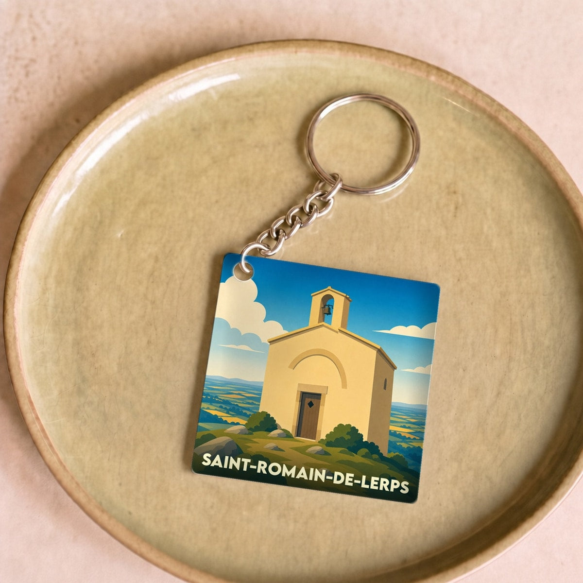 Porte-clés souvenir de la ville de Saint-Romain-de-Lerps avec une illustration stylisée d'une chapelle sous un ciel bleu.