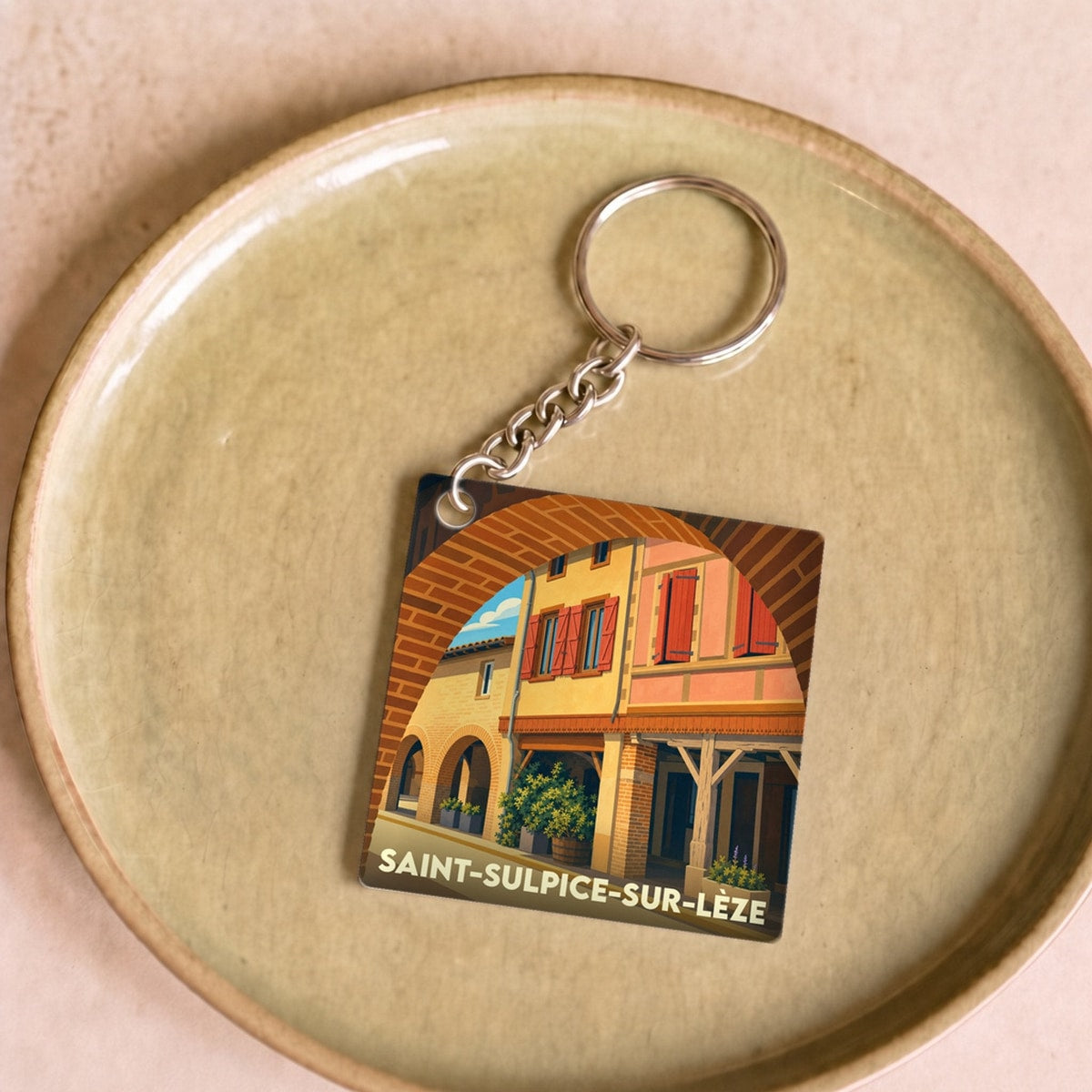Porte-clés souvenir de la ville de Saint-Sulpice-sur-Lèze avec une illustration stylisée d'une rue pittoresque sous une arche.