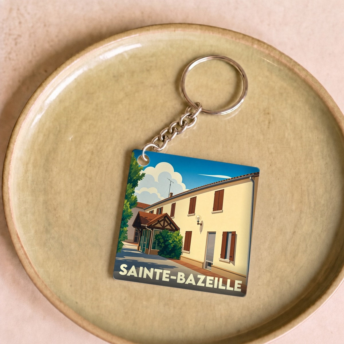 Porte-clés souvenir de la ville de Sainte-Bazeille avec une illustration stylisée d'un bâtiment ensoleillé.