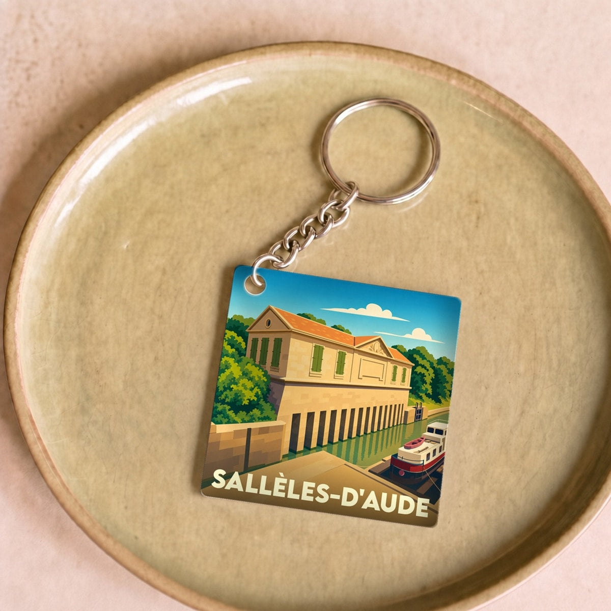 Porte-clés souvenir de la ville de Sallèles-d'Aude avec une illustration stylisée d'un bâtiment au bord de l'eau.