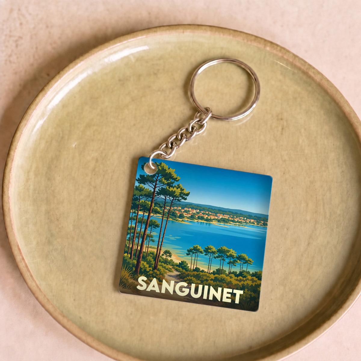 Porte-clés souvenir de la ville de Sanguinet avec une illustration stylisée d'un paysage côtier bordé de pins.