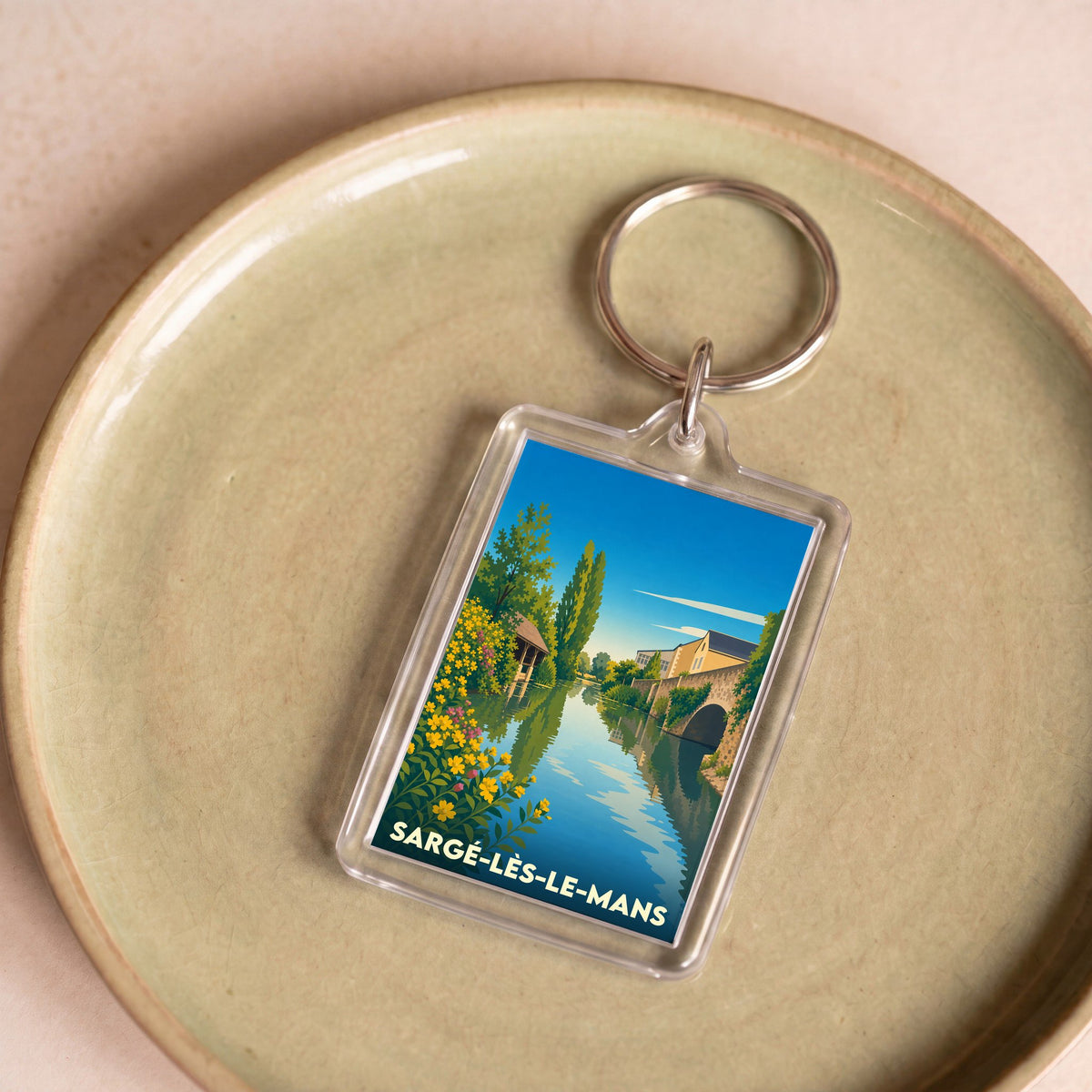 Porte-clés souvenir de Sargé-lès-le-Mans avec une illustration stylisée d'un cours d'eau bordé de fleurs et d'un pont.