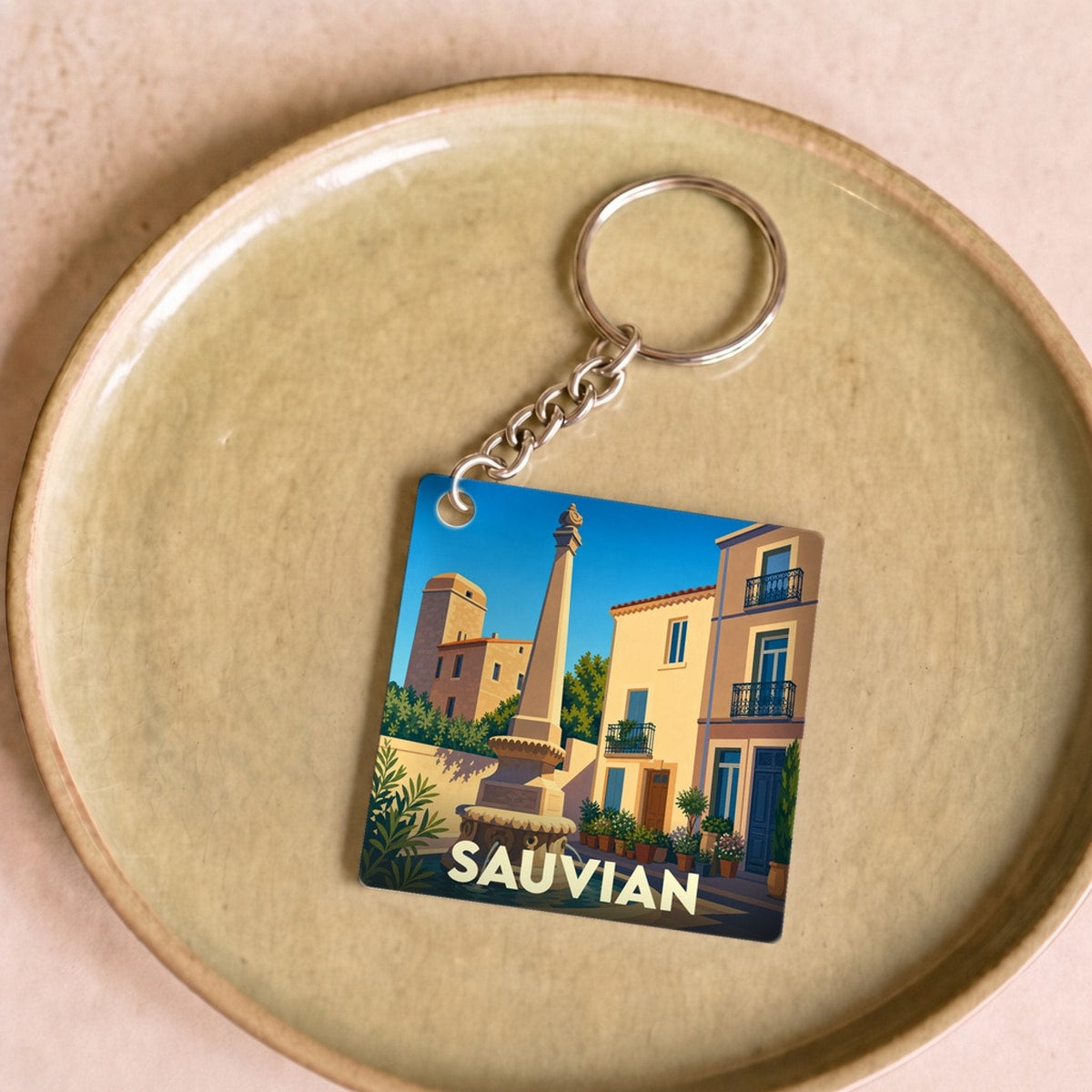 Porte-clés souvenir de la ville de Sauvian avec une illustration stylisée d'une place ensoleillée et sa fontaine.