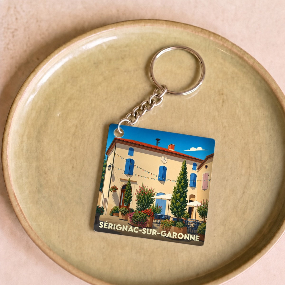 Porte-clés souvenir de Sérignac-sur-Garonne avec une illustration stylisée d'une place ensoleillée.