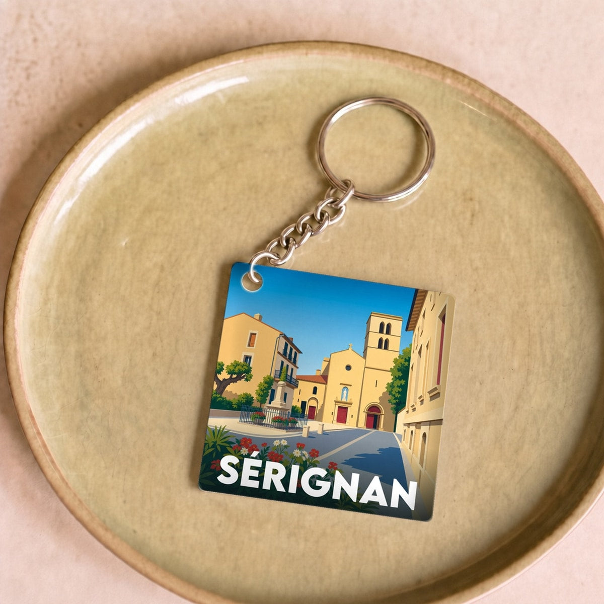 Porte-clés souvenir de la ville de Sérignan avec une illustration stylisée d'une place ensoleillée.