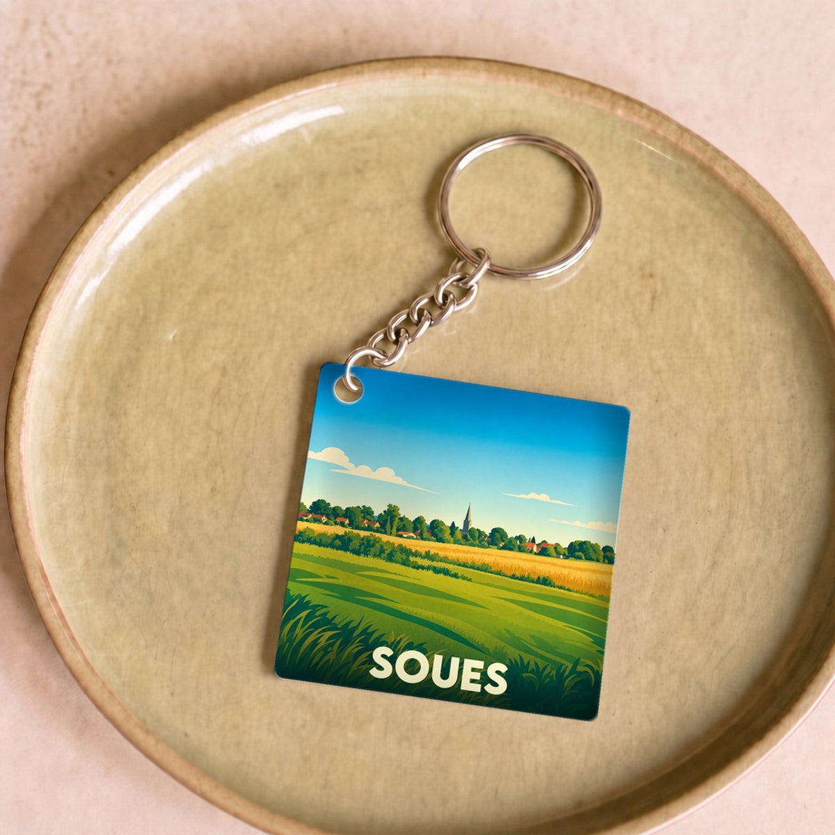 Porte-clés souvenir de la ville de Soues avec une illustration stylisée de paysages champêtres.