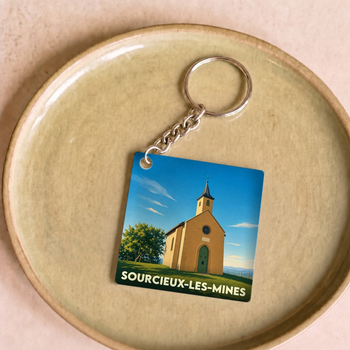 Porte-clés souvenir de la ville de Sourcieux-les-Mines avec une illustration stylisée d'une chapelle sous un ciel bleu.