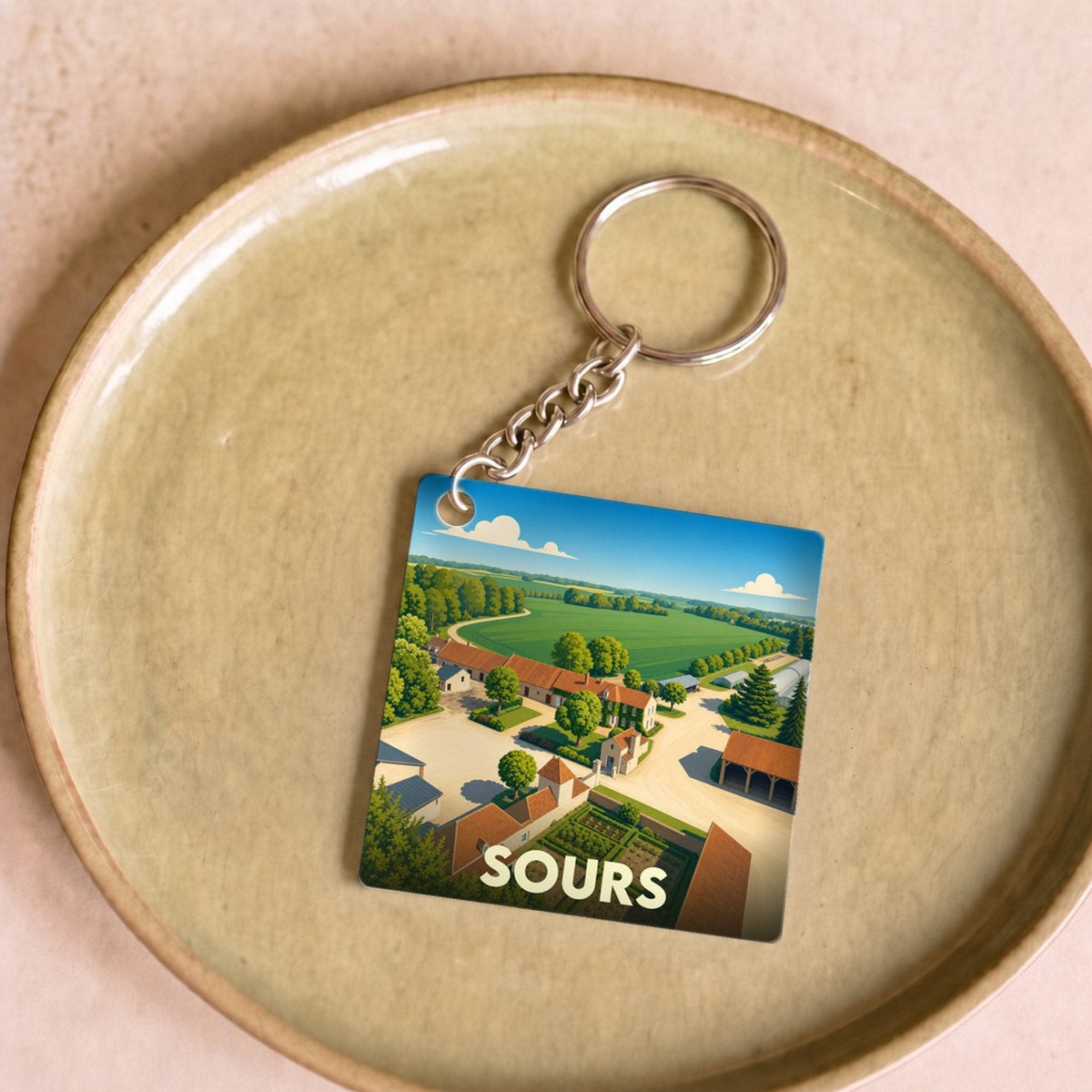 Porte-clés souvenir de la ville de Sours avec une illustration stylisée de ses paysages ruraux.