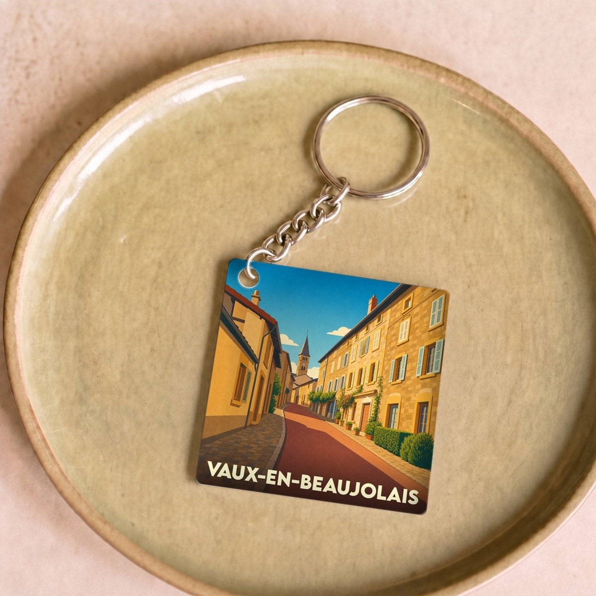 Porte-clés souvenir de la ville de Vaux-en-Beaujolais avec une illustration stylisée d'une rue ensoleillée.
