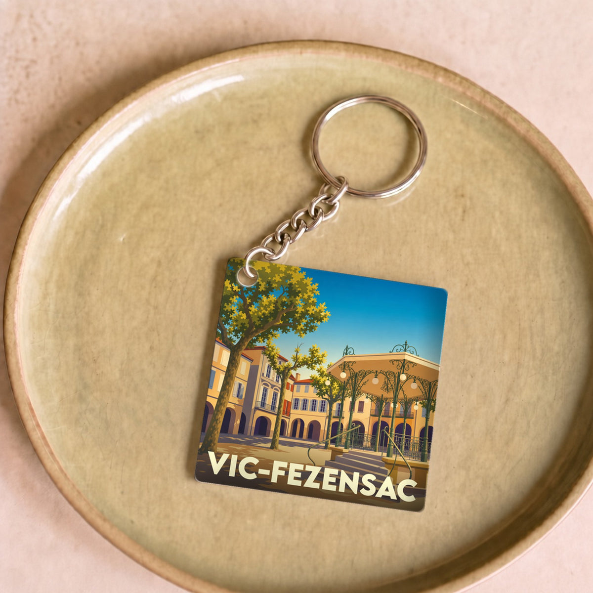 Porte-clés souvenir de la ville de Vic-Fezensac avec une illustration stylisée d'un kiosque et d'une place ensoleillée.