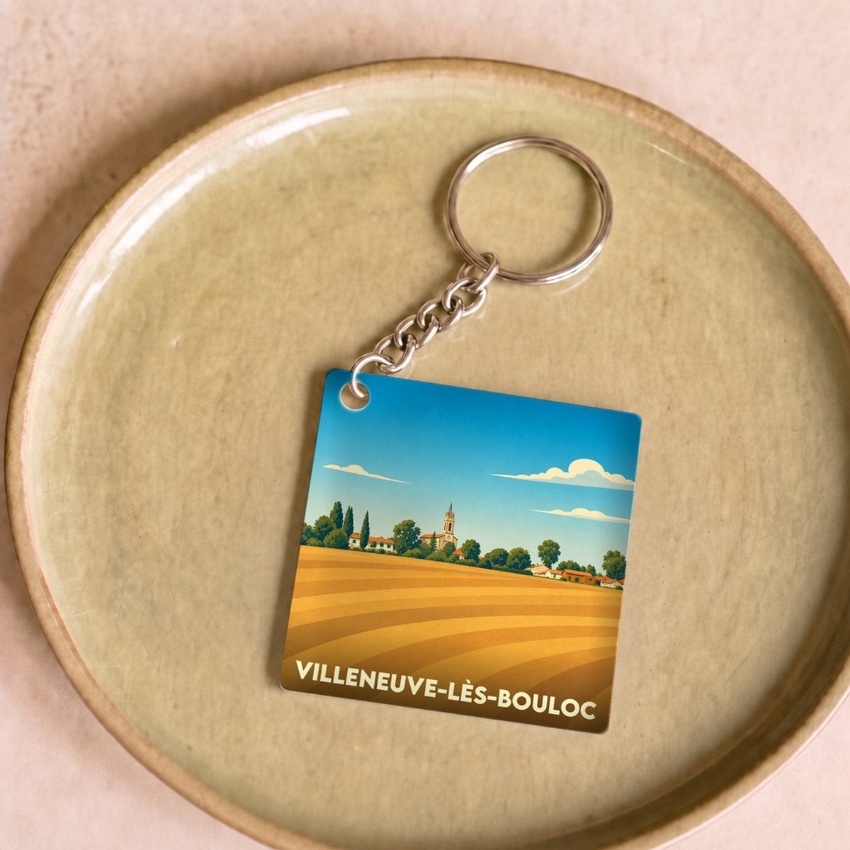Porte-clés souvenir de la ville de Villeneuve-lès-Bouloc avec une illustration stylisée d'un paysage de champs.