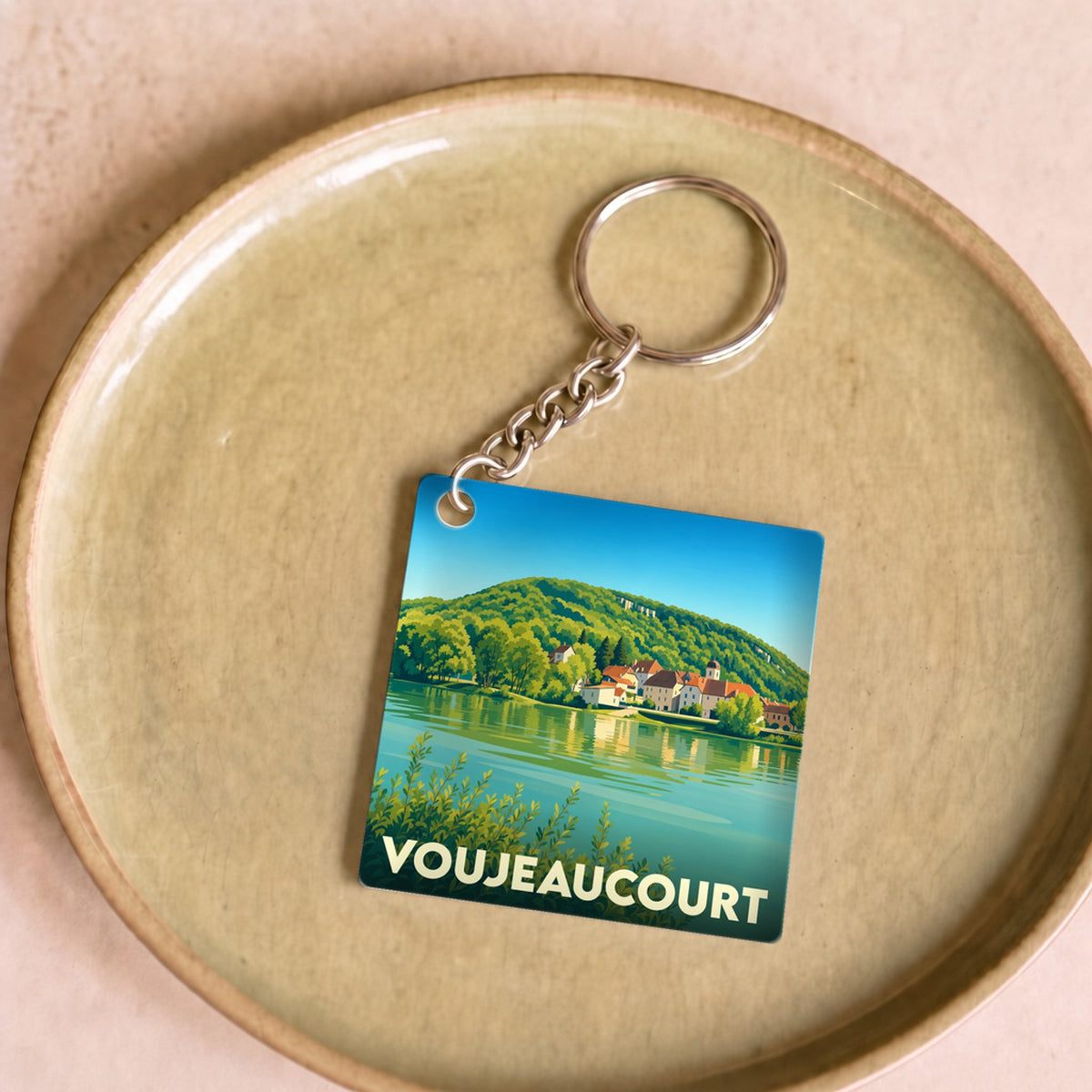 Porte-clés souvenir de la ville de Voujeaucourt avec une illustration stylisée d'un paysage de bord de rivière.