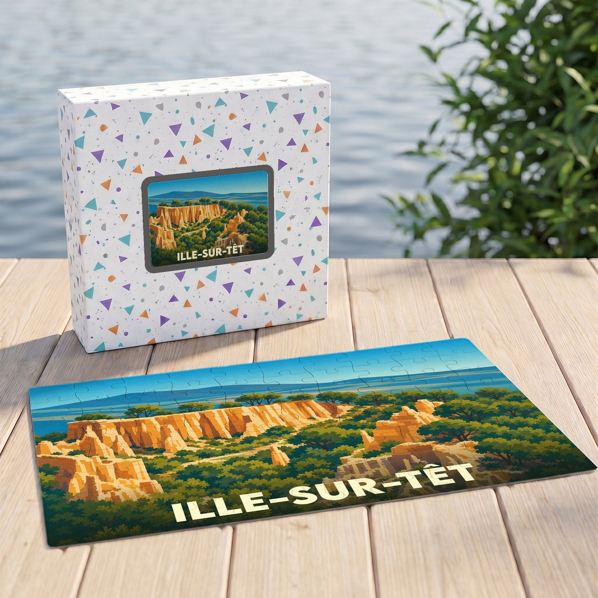 Puzzle souvenir d'Ille-sur-Têt présentant une illustration stylisée des Orgues, posé à côté de sa boîte sur un ponton en bois.