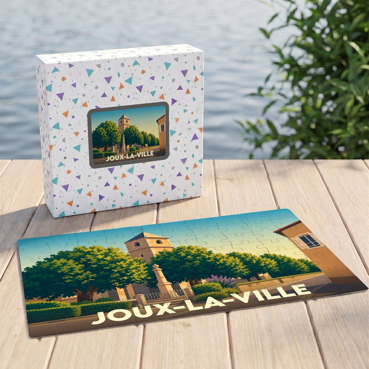 Puzzle souvenir de la ville de Joux-la-Ville avec son coffret, illustrant une place arborée et son église dans un style graphique épuré.