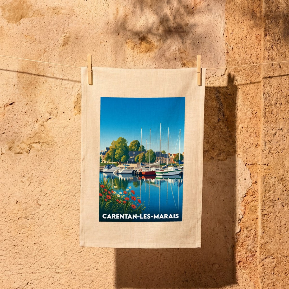 Torchon souvenir de la ville de Carentan-les-Marais illustrant un port de plaisance avec des voiliers et des fleurs colorées au premier plan.