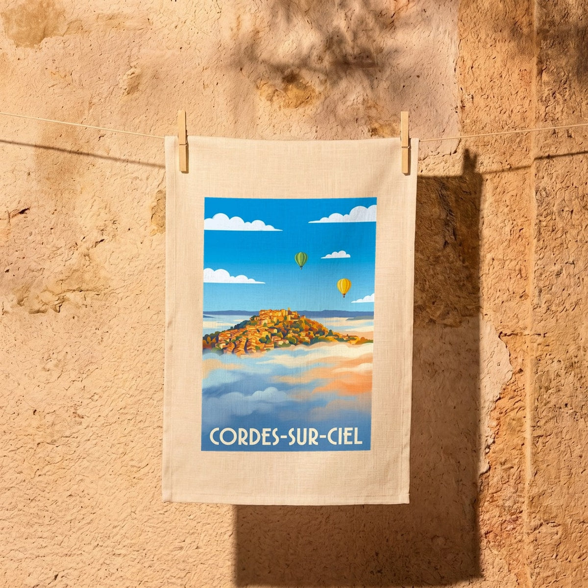 Torchon souvenir de Cordes-sur-Ciel illustré d'une vue stylisée du village émergeant des nuages avec des montgolfières.