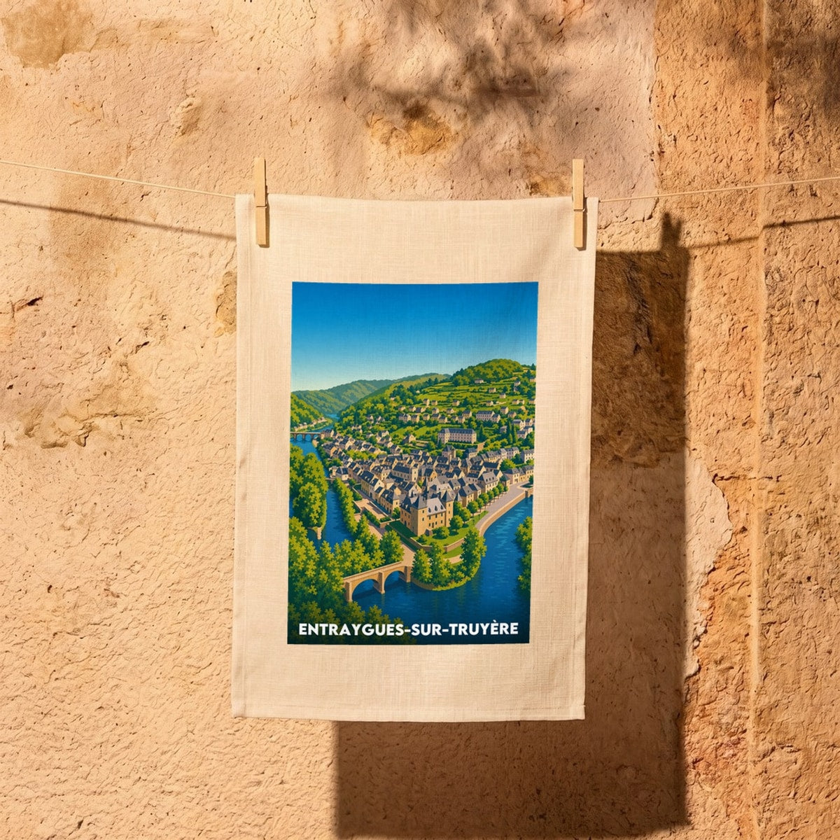 Torchon souvenir de la ville d’Entraygues-sur-Truyère avec une illustration stylisée et colorée du village bordé par l'eau.