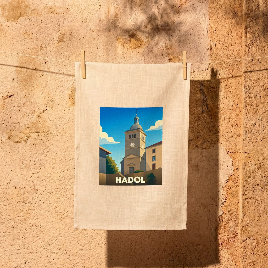 Torchon souvenir de la ville de Hadol illustré d'une église sous un ciel bleu, suspendu par des pinces à linge devant un mur en pierre.