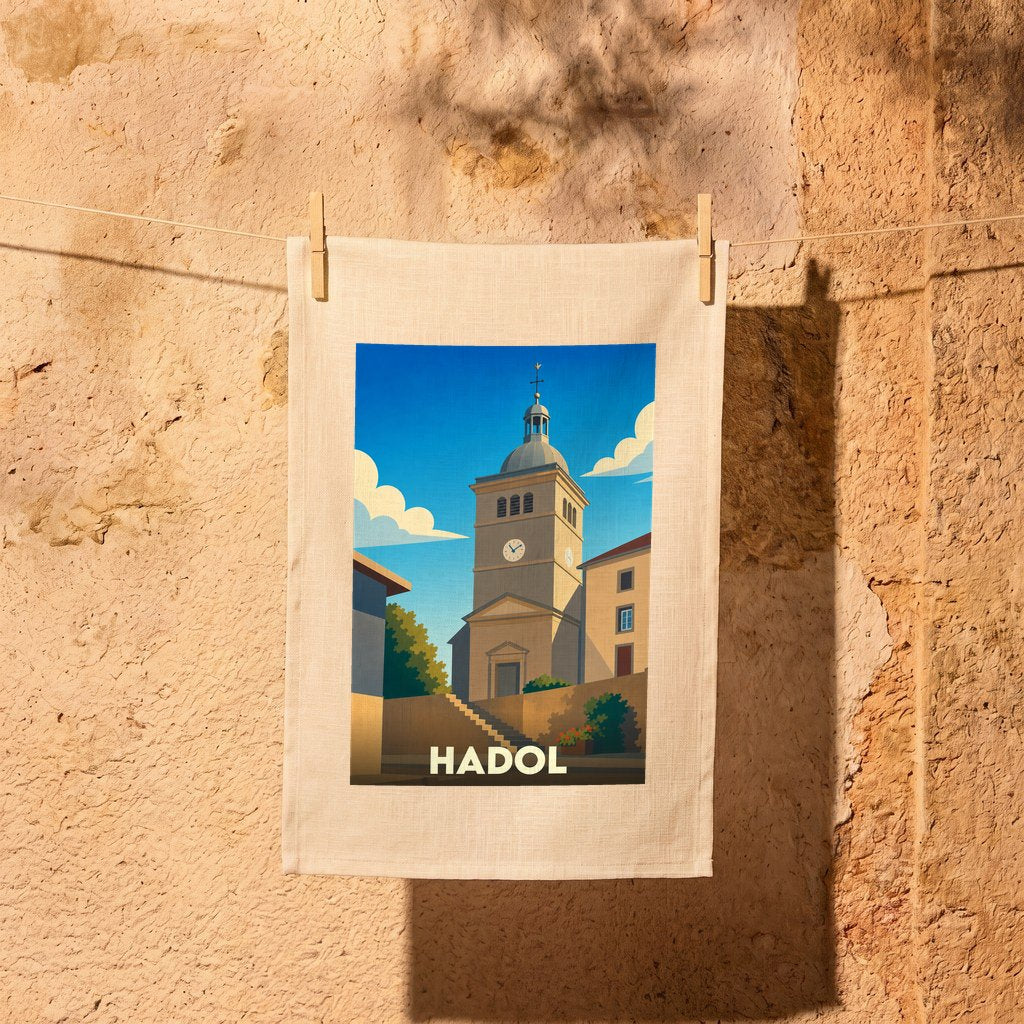 Torchon souvenir de la ville de Hadol illustré d'une église sous un ciel bleu, suspendu par des pinces à linge devant un mur en pierre.