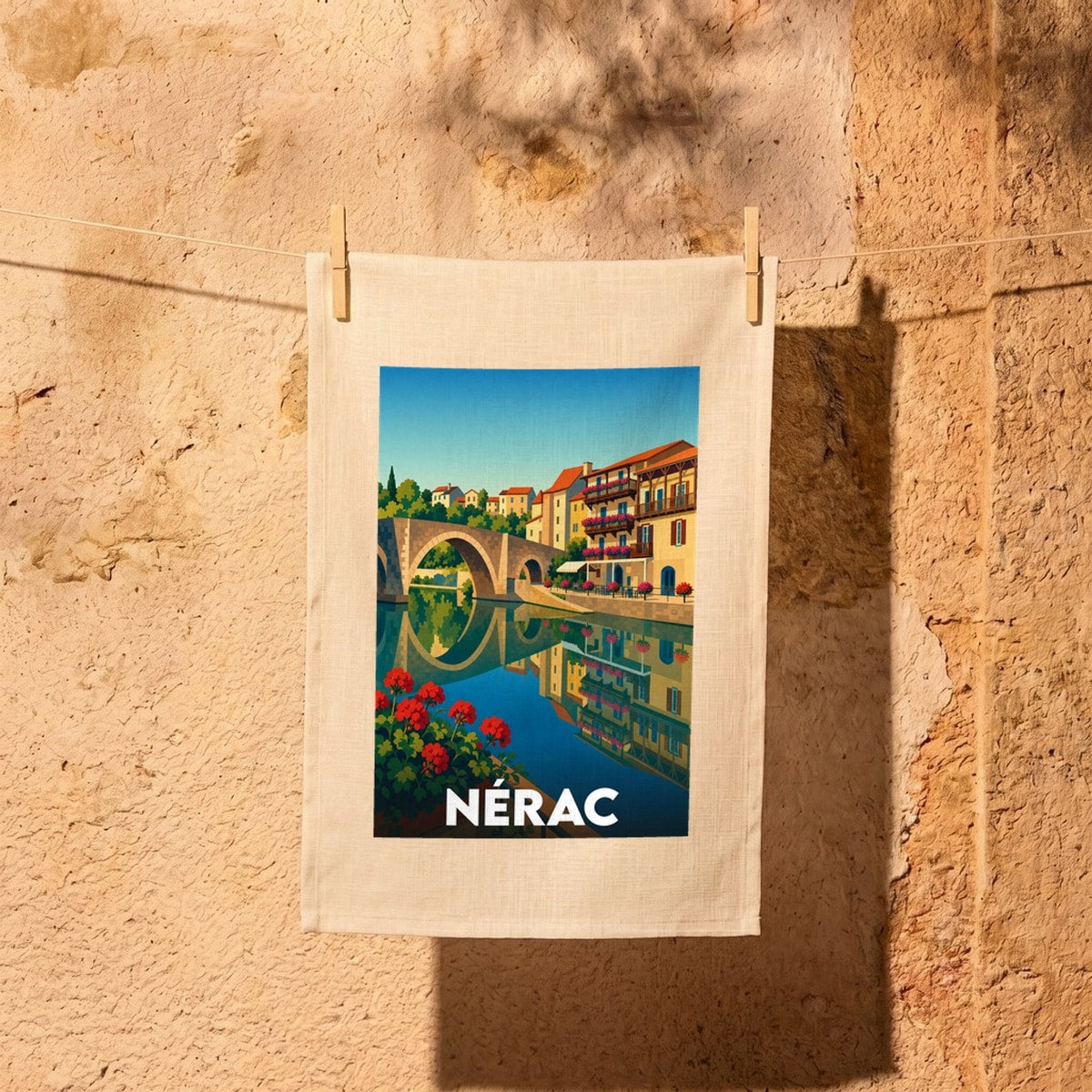 Torchon souvenir de la ville de Nérac orné d'une illustration stylisée représentant son pont vieux et ses maisons à colombages au bord de l'eau.
