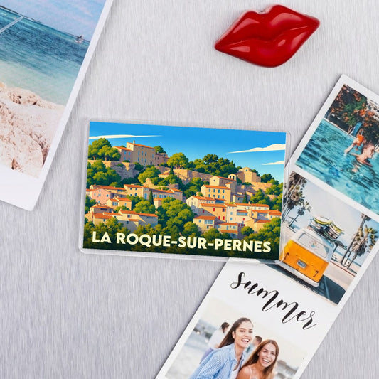 Magnet souvenir de la ville de La Roque-sur-Pernes avec une illustration colorée et stylisée du village.