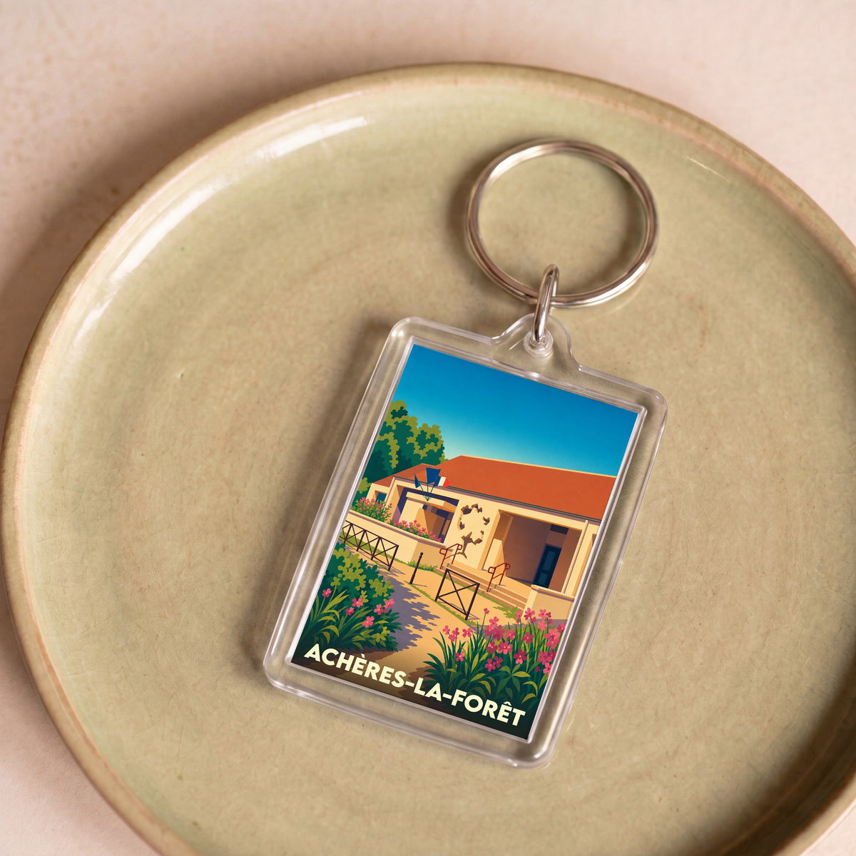 Porte-clés souvenir de la ville d'Achères-la-Forêt avec une illustration stylisée d'un bâtiment fleuri.