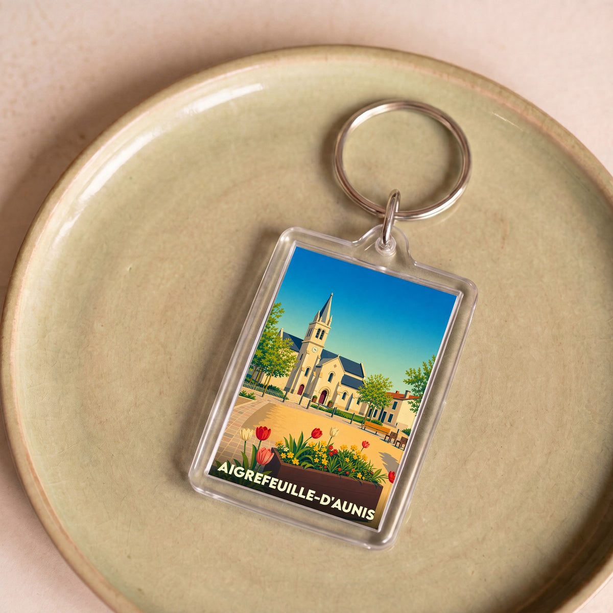 Porte-clés souvenir de la ville d'Aigrefeuille-d'Aunis avec une illustration stylisée de son église et de fleurs colorées.