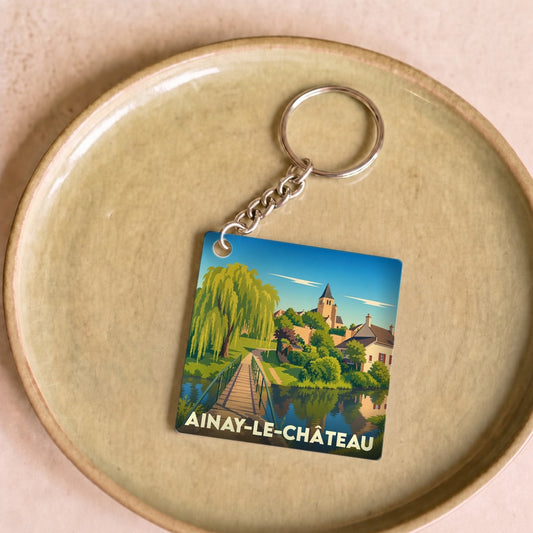 Porte-clés souvenir de la ville d'Ainay-le-Château avec une illustration stylisée d'un paysage fluvial.