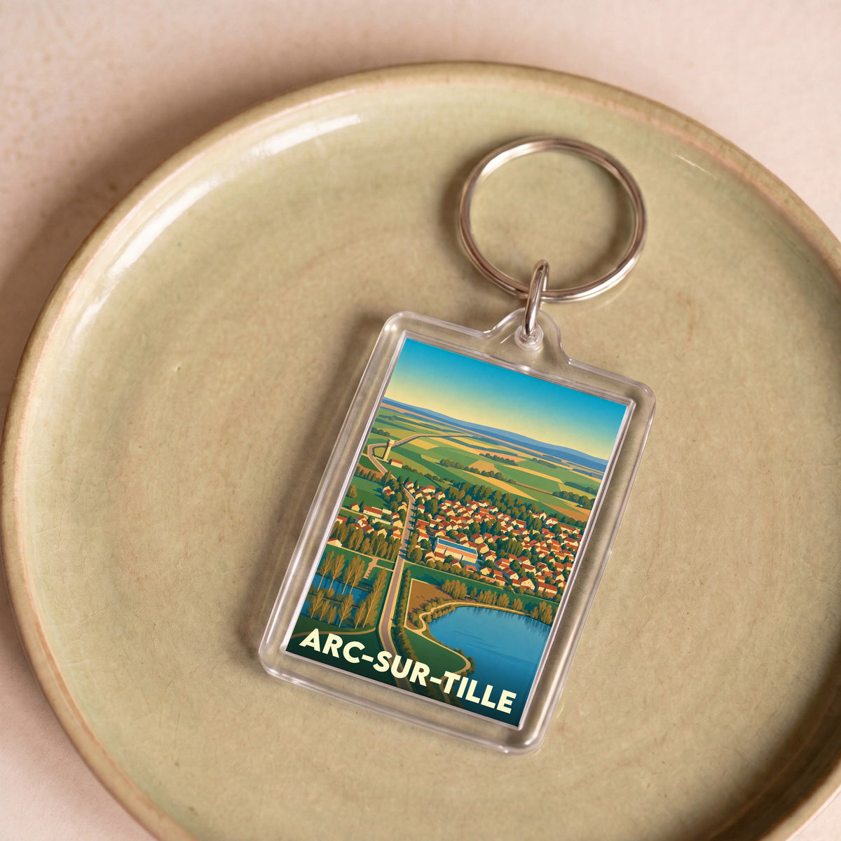 Porte-clés souvenir de la ville d'Arc-sur-Tille avec une illustration stylisée d'un paysage aérien.