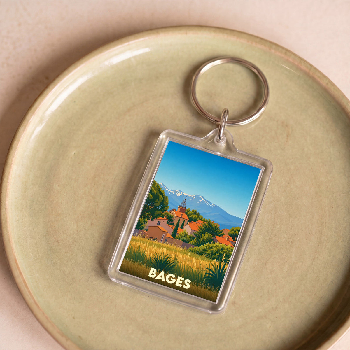 Porte-clés souvenir de la ville de Bages avec une illustration stylisée d'un village face aux montagnes.