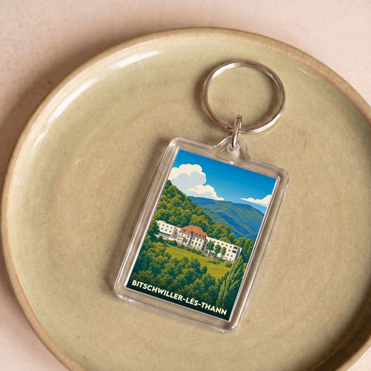Porte-clés souvenir de la ville de Bitschwiller-lès-Thann avec une illustration stylisée d'un bâtiment dans un paysage de montagne.
