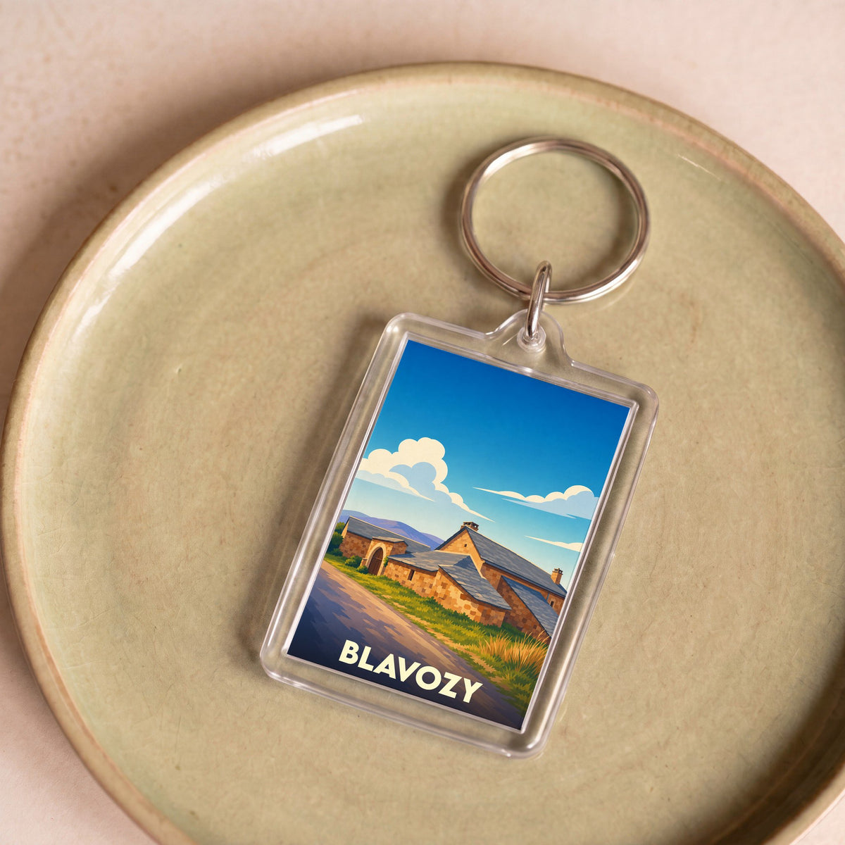 Porte-clés souvenir de la ville de Blavozy avec une illustration stylisée de maisons en pierre sous un ciel bleu.