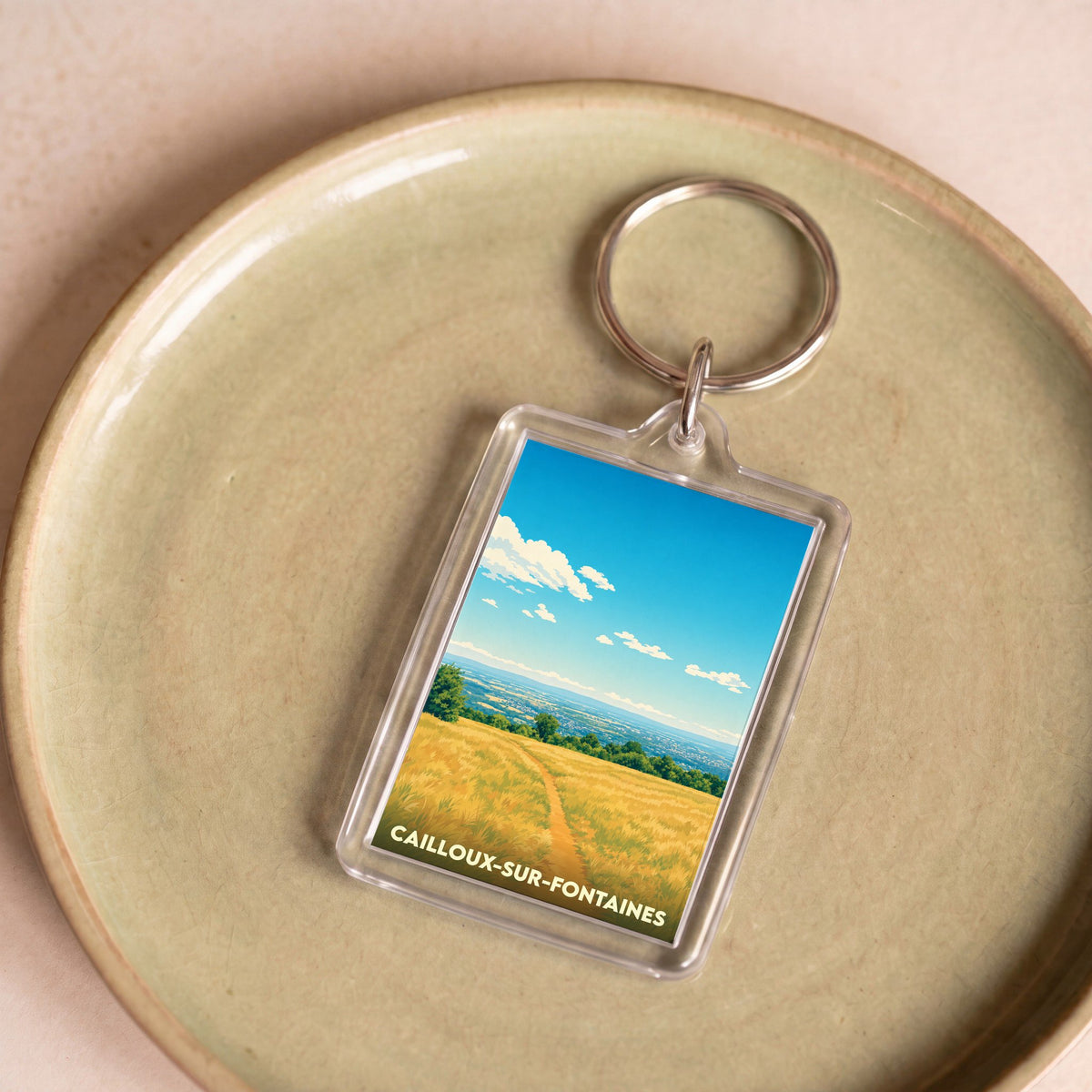 Porte-clés souvenir de la ville de Cailloux-sur-Fontaines avec une illustration stylisée d'un paysage champêtre.